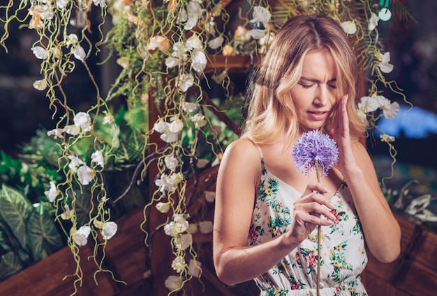 Verwirrte blonde junge Frau, die in der Hand künstliche purpurrote Lauchblume hält