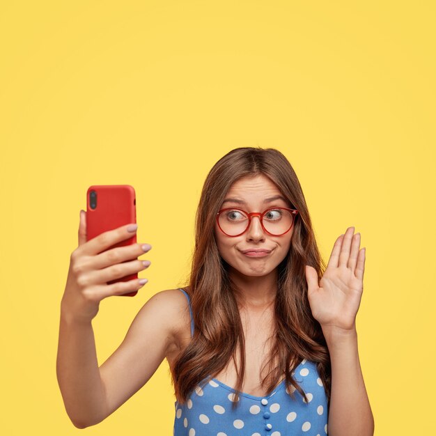 Verwirrte brünette Frau begrüßt Freund während des Videoanrufs, winkt mit der Handfläche an der Kamera, hält modernes Handy vor Gesicht
