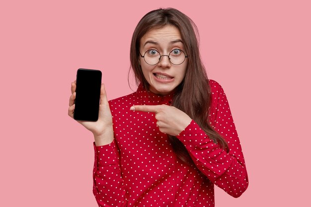 Verwirrte dunkelhaarige junge Frau in optischer Brille, zeigt auf elektronisches Gerät mit Modellbildschirm, trägt rotes Hemd, wirbt für neues Gerät, hat grüne Augen