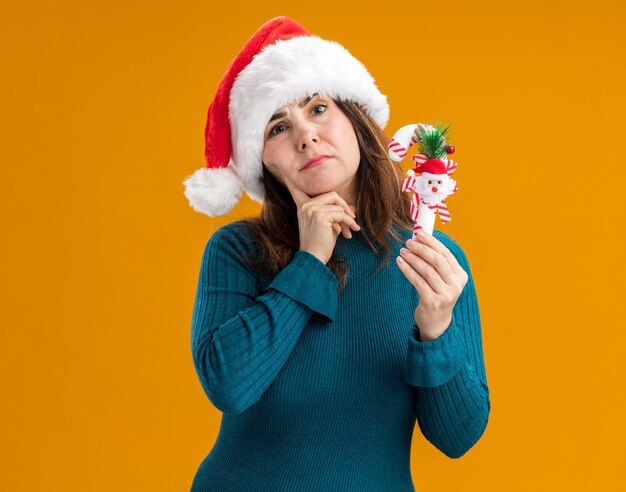 Verwirrte erwachsene kaukasische Frau mit Weihnachtsmütze legt Finger auf Kinn und hält Zuckerstange lokalisiert auf orange Hintergrund mit Kopienraum