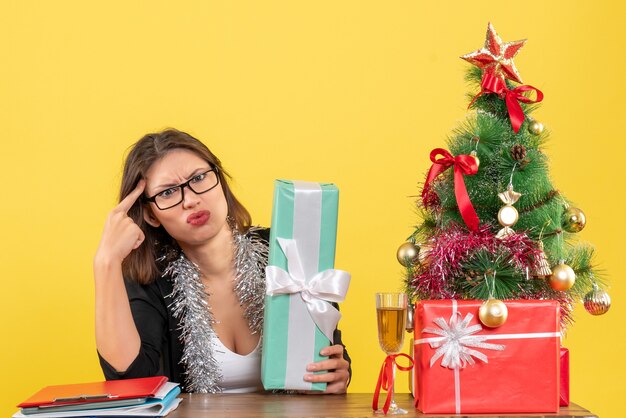 Verwirrte Geschäftsdame im Anzug mit Brille, die ihr Geschenk zeigt und an einem Tisch mit einem Weihnachtsbaum darauf im Büro sitzt