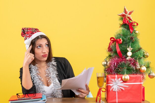 Verwirrte Geschäftsdame im Anzug mit Weihnachtsmannhut und Neujahrsdekorationen, die Dokumente halten und an einem Tisch mit einem Weihnachtsbaum darauf im Büro sitzen