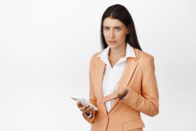 Verwirrte Geschäftsfrau, die auf ihr Handy zeigt und verwirrt in die Kamera starrt, die im Anzug vor weißem Hintergrund steht