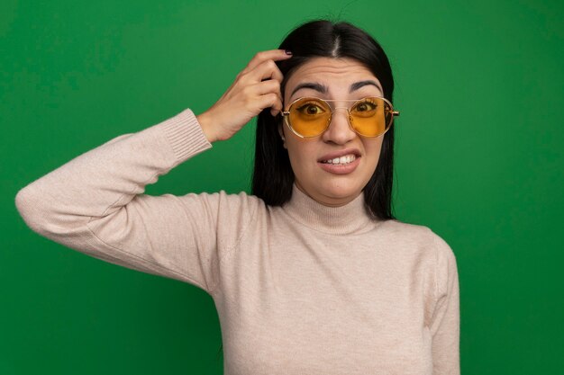 Verwirrte hübsche brünette Frau in Sonnenbrille setzt Finger auf Kopf isoliert auf grüner Wand