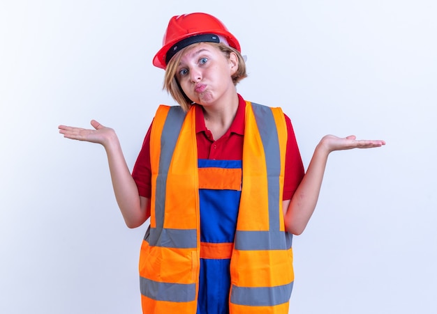 Kostenloses Foto verwirrte junge baumeisterin in uniform, die hände isoliert auf weißer wand ausbreitet