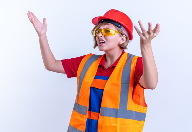 Verwirrte junge Baumeisterin in Uniform mit Brille, die die Hände isoliert auf weißer Wand hebt