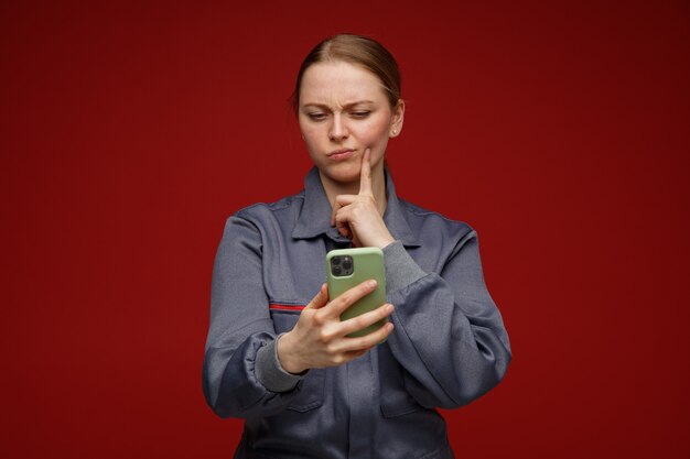 Verwirrte junge blonde Ingenieurin, die Uniform trägt, die das Gesicht des Fingers mit dem Finger hält und betrachtet