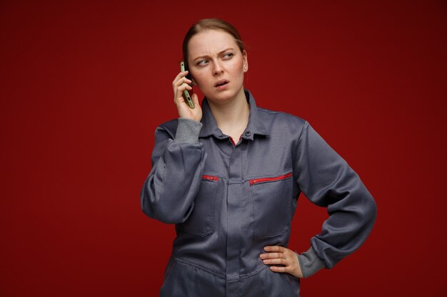 Verwirrte junge blonde Ingenieurin in Uniform, die am Telefon spricht und die Hand auf der Taille hält und die Seite betrachtet