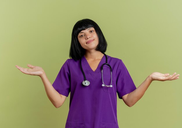 Verwirrte junge brünette Ärztin in Uniform mit Stethoskop steht mit offenen Händen