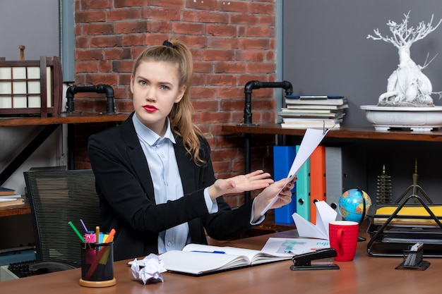 Verwirrte junge Dame, die an einem Tisch sitzt und das Dokument im Büro zeigt