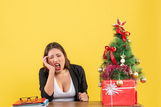 Verwirrte junge Frau, die an einem Tisch im Anzug nahe geschmücktem Weihnachtsbaum im Büro auf lokalisiertem Gelb sitzt