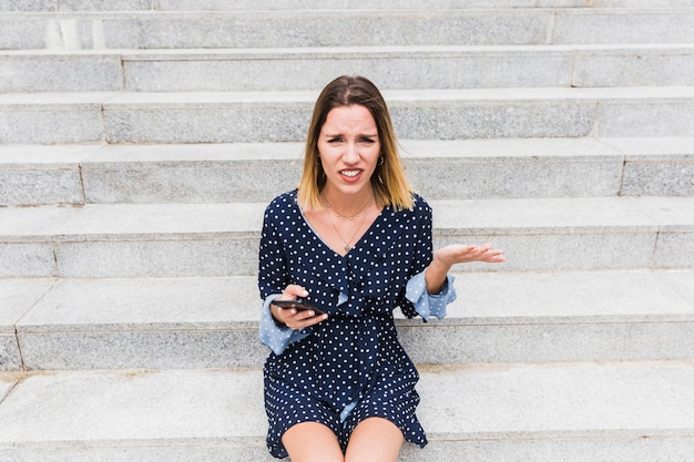 Verwirrte junge Frau, die auf dem Treppenhaus hält Smartphone sitzt