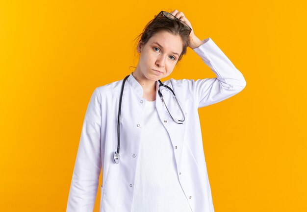 Verwirrte junge Frau in Arztuniform mit Stethoskop am Kopf kratzen