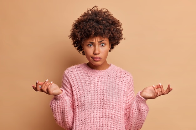 Kostenloses Foto verwirrte junge frau mit afro-haarschnitt spreizt handflächen und steht vor dilemma hat schwierige wahl in lässigen strickpullover über braune wand isoliert gekleidet