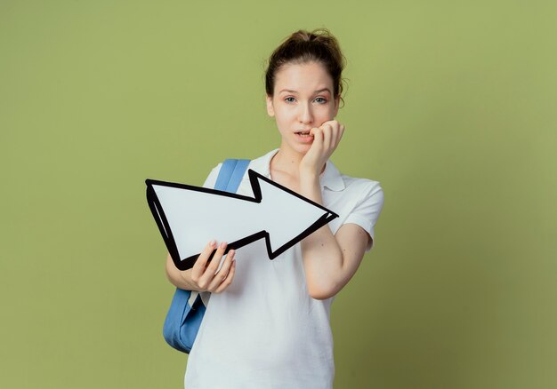 Verwirrte junge hübsche Studentin, die Rückentasche hält, die Pfeilmarkierung hält, die zur Seite zeigt und Hand auf Lippe lokalisiert auf olivgrünem Hintergrund mit Kopienraum legt