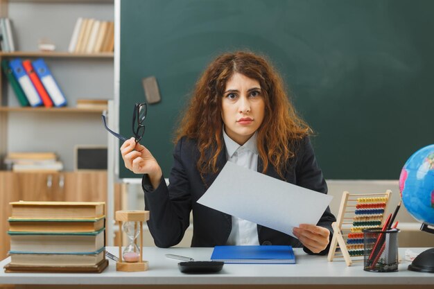 verwirrte junge lehrerin, die papier mit brille trägt und mit schulwerkzeugen im klassenzimmer am schreibtisch sitzt