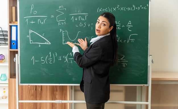 Verwirrte junge Lehrerin, die vor der Tafel steht, zeigt an der Tafel im Klassenzimmer