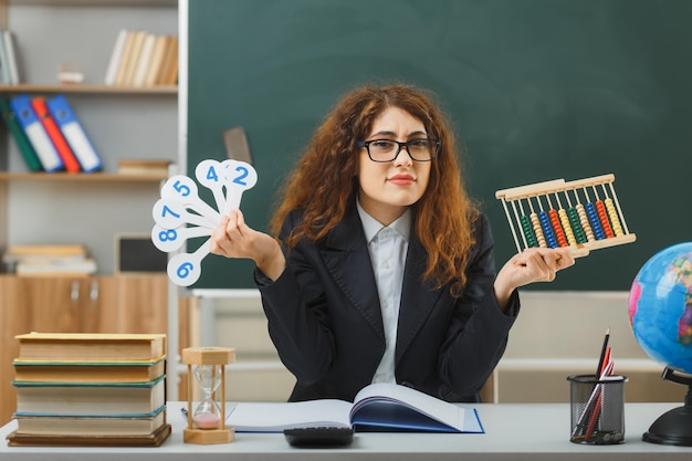 Kostenloses Foto verwirrte junge lehrerin mit brille, die abakus mit zahlenfächern hält, die am schreibtisch mit schulwerkzeugen im klassenzimmer sitzen