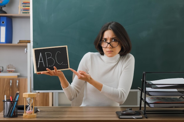 verwirrte junge lehrerin mit brille und zeigt auf eine mini-tafel, die mit schulwerkzeugen im klassenzimmer am schreibtisch sitzt