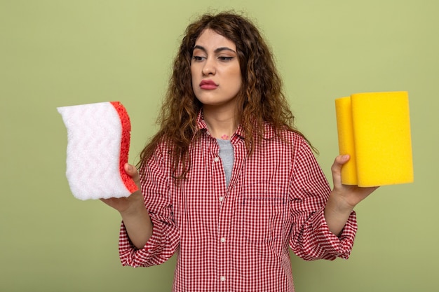 Kostenloses Foto verwirrte junge putzfrau, die reinigungsschwämme hält und betrachtet