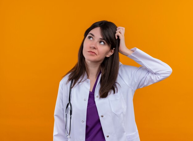 Verwirrte junge Ärztin im medizinischen Gewand mit Stethoskop legt Hand auf Kopf auf lokalisierten orange Hintergrund mit Kopienraum