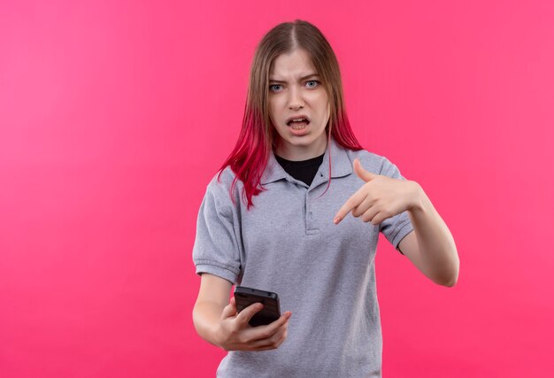 Verwirrte junge schöne Frau, die graues T-Shirt trägt, zeigt auf Telefon in ihrer Hand auf isolierter rosa Wand