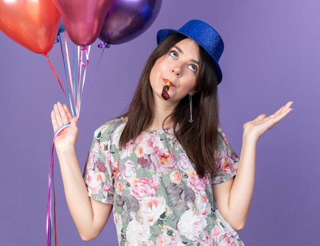 Verwirrte junge schöne Frau mit Partyhut, die Luftballons hält und die Partypfeife bläst, die Hände isoliert auf blauer Wand ausbreitet