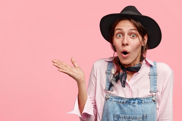 Verwirrte schöne Frau mit zögerndem Gesichtsausdruck, öffnet den Mund, trägt ein lässiges Outfit, hebt die Handfläche, zeigt einen seltsamen Ort, posiert an der rosa Wand