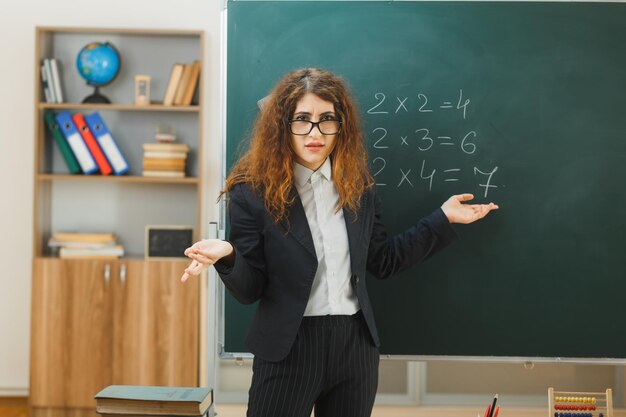 Verwirrte sich ausbreitende Hände Junge Lehrerin, die vor der Tafel steht und im Klassenzimmer zeigt