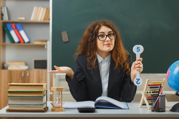 verwirrte sich ausbreitende hände junge lehrerin mit brille, die einen fächer hält, der am schreibtisch mit schulwerkzeugen im klassenzimmer sitzt