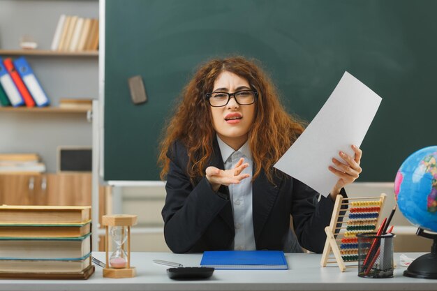 Verwirrte sich ausbreitende Hand Junge Lehrerin mit Brille, die Papier hält und am Schreibtisch mit Schulwerkzeugen im Klassenzimmer sitzt