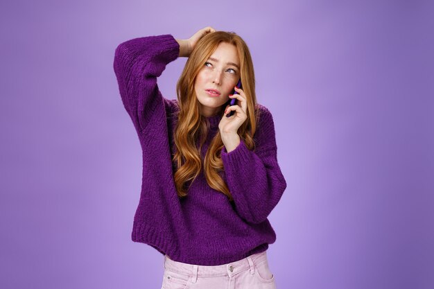 Verwirrte süße Rothaarigefrau, die per Handy eine Verabredung macht, den Hinterkopf kratzt und schielt, aufschaut, als denkend, eine Wahl treffen oder sich erinnern, mit dem Smartphone über die violette Wand sprechen.