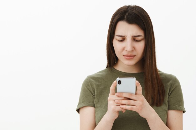 Verwirrte und frustrierte junge Frau kann nicht verstehen, was es bedeutet, wenn eine Nachricht die Stirn runzelt und auf dem Smartphone-Bildschirm befragt aussieht, als würde sie einen Strage-Text lesen, ahnungslos posieren und sich über die graue Wand konzentrieren
