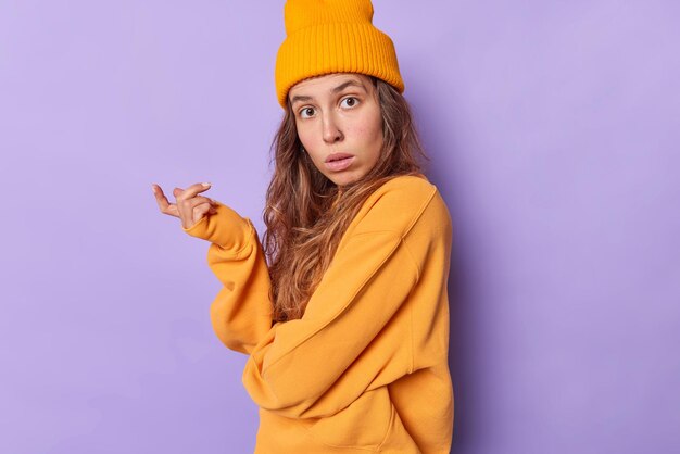 Verwirrte, zögernde Frau mit langen Haaren hat einen empörten Ausdruck und sagt, dass das, was einen orangefarbenen Hut und einen losen Pullover trägt, seitlich vor violettem Hintergrund steht, nicht verstehen kann, was Sie von ihr wollen.