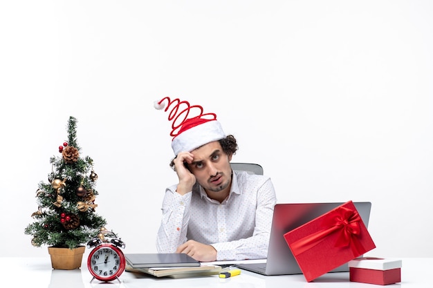 Verwirrter beschäftigter junger Geschäftsmann mit lustigem Weihnachtsmannhut, der an etwas im Büro auf weißem Hintergrund denkt