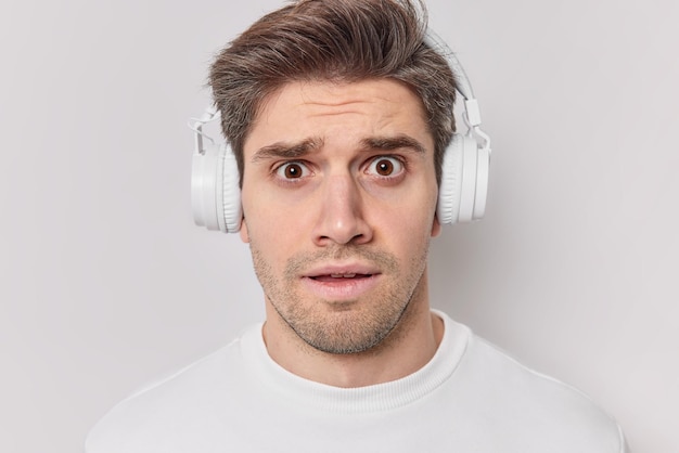 Verwirrter besorgter männlicher Student mit dunklem Haar hört Vorlesung über Kopfhörer zu und versucht, sich an Informationen für die Prüfung zu erinnern, sieht belästigt gekleidet aus, lässig isoliert vor weißem Hintergrund. Menschliche Reaktionen
