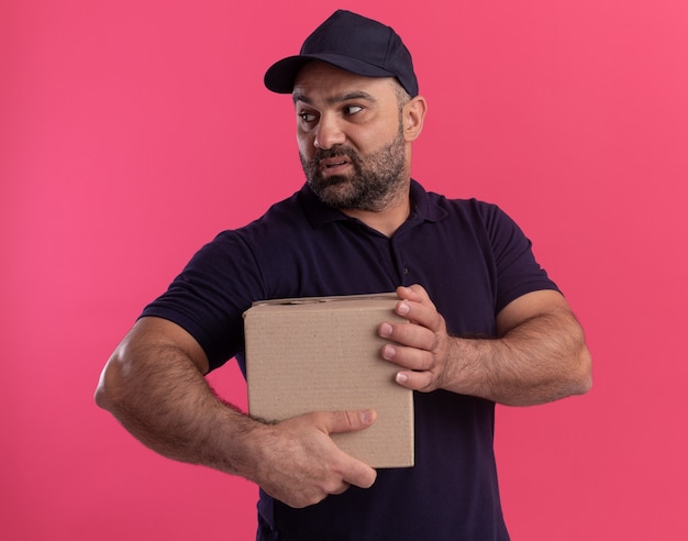 Verwirrter Blick auf den seitlichen Lieferboten mittleren Alters in Uniform und Kappenhaltebox isoliert auf rosa Wand