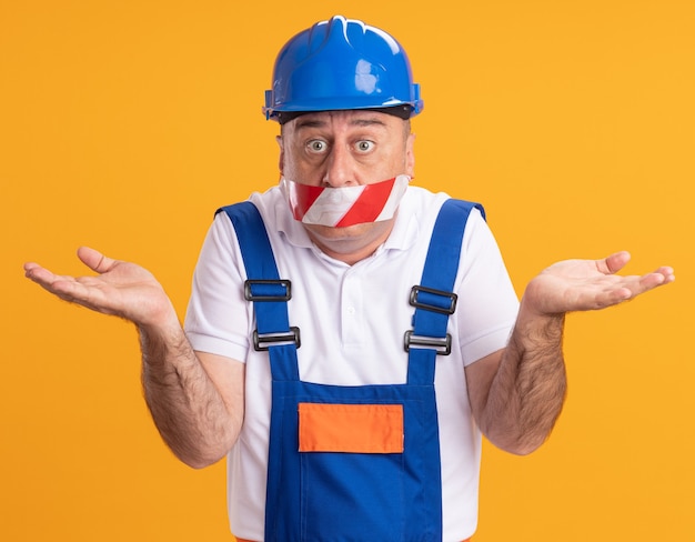 Kostenloses Foto verwirrter erwachsener baumeister in uniform bedeckt mund mit klebeband und hält hände offen auf orange wand offen