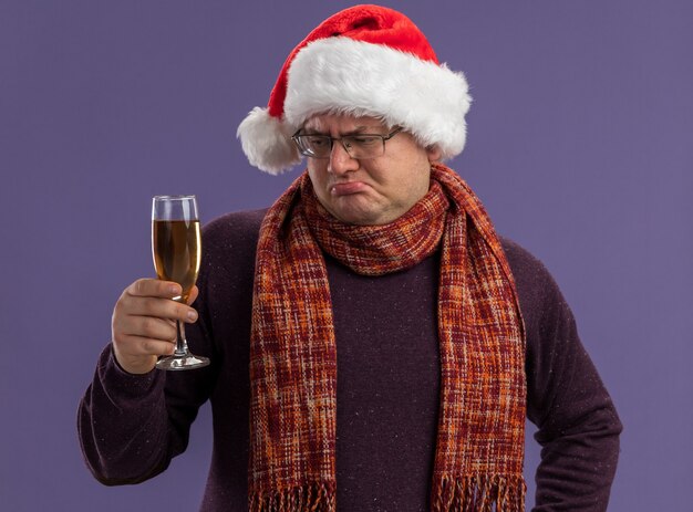 verwirrter erwachsener Mann, der Brille und Weihnachtsmütze mit Schal um Hals hält und Glas Champagner auf lila Hintergrund isoliert betrachtet