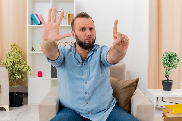 Verwirrter erwachsener slawischer Mann sitzt auf Sessel und gestikuliert sechs mit den Fingern im Wohnzimmer