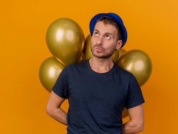 Verwirrter hübscher kaukasischer Mann, der blauen Parteihut trägt, hält Luftballons hinter lokalisiert auf orange Hintergrund mit Kopienraum