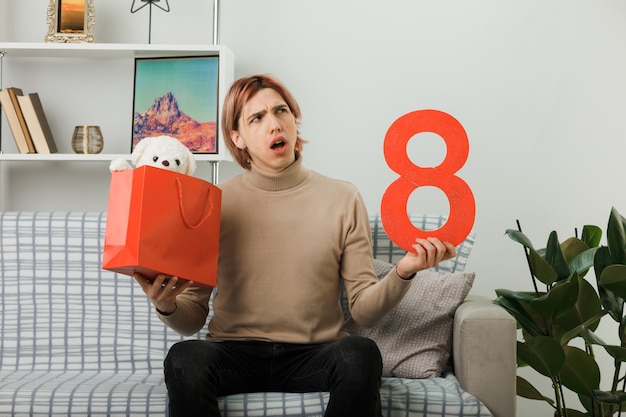Verwirrter hübscher Kerl am glücklichen Frauentag, der die Nummer acht mit Geschenktüte auf dem Sofa im Wohnzimmer hält