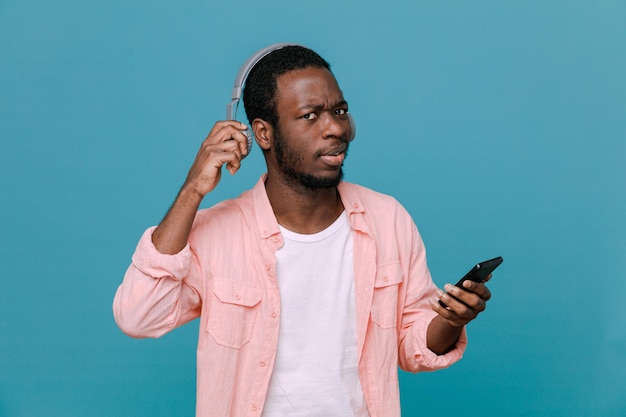 Verwirrter junger Afroamerikaner, der das Telefon mit Kopfhörern isoliert auf blauem Hintergrund hält