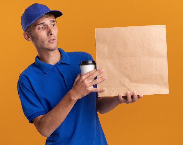 Verwirrter junger blonder Lieferjunge hält eine Tasse zum Mitnehmen und sieht sich das Papierpaket an, das auf einer orangefarbenen Wand mit Kopierraum isoliert ist