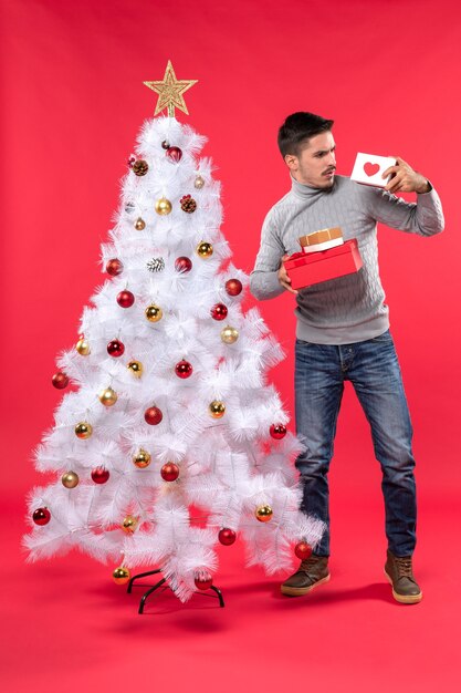 Verwirrter junger Erwachsener in einer grauen Bluse, die nahe dem geschmückten Weihnachtsbaum steht und seine Geschenke auf Rot hält