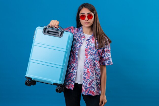 Verwirrter junger Frauentourist, der rote Sonnenbrille hält, die Reisekoffer mit verärgertem Gesichtsausdruck hält, der auf lokalisiertem Blau steht