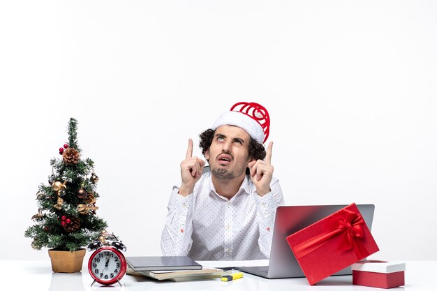 Verwirrter junger Geschäftsmann mit lustigem Weihnachtsmannhut, der an etwas denkt, indem er oben im Büro auf weißem Hintergrund schaut