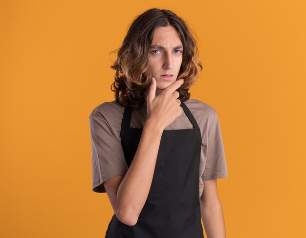 Verwirrter junger gutaussehender Friseur in Uniform, der die Hand am Kinn hält, isoliert auf oranger Wand mit Kopierraum