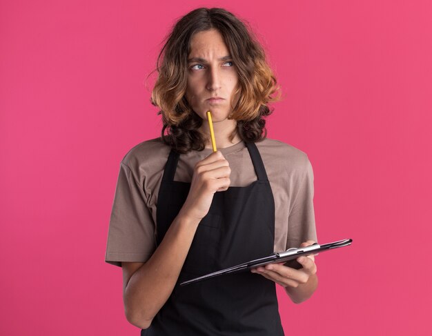 Verwirrter junger gutaussehender Friseur in Uniform, der die Zwischenablage an der Seite betrachtet und das Kinn mit einem Bleistift berührt