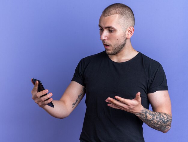 Kostenloses Foto verwirrter junger gutaussehender kerl mit schwarzem t-shirt, das telefon auf blauem hintergrund hält und sieht